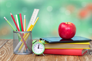 stock photo of pencils, books, laptop, apple, clock on desk in classroom.